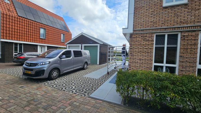 Een grijze bestelwagen naast een garage in een woonomgeving met stenen en gras.